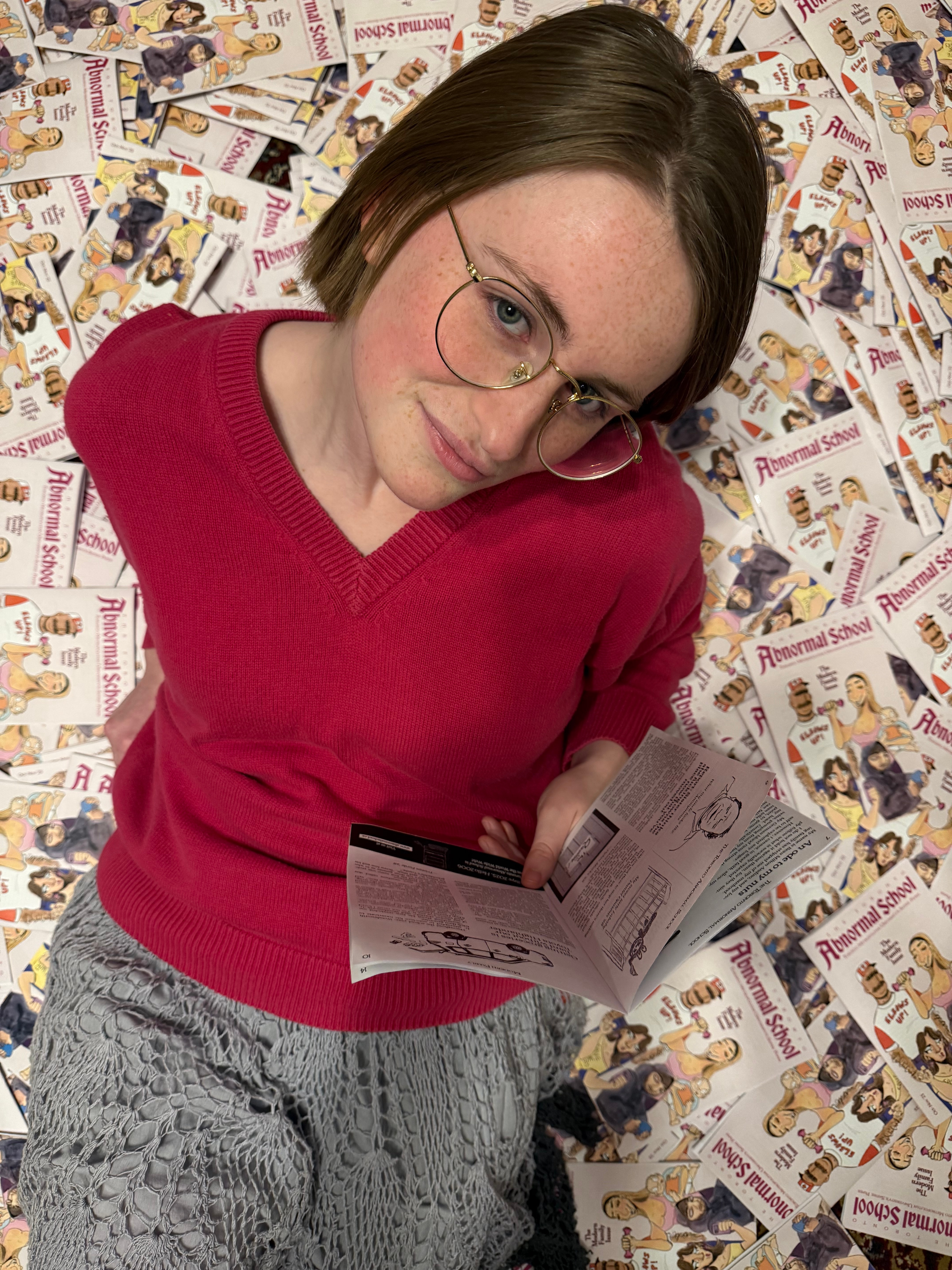 A photograph of a young woman wearing a pink sweater and grey skirt laying on a pile of magazines. She is wearing gold rimmed glasses and holding a magazine in her left hand. 